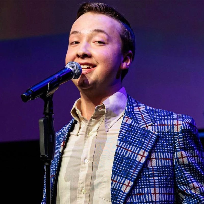 A young man with short brown hair smiles while singing into a microphone. He wears a white dress shirt and a blue, red, and white checkered blazer. The background is dimly lit with purple and blue tones, suggesting a stage setting.