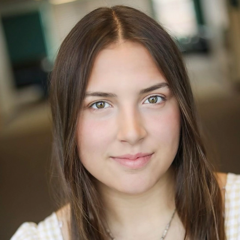 A young woman with long straight brown hair and fair skin looks directly at the camera, softly smiling. She wears a light top and a necklace. The background is blurred, suggesting an indoor setting with green and white tones.