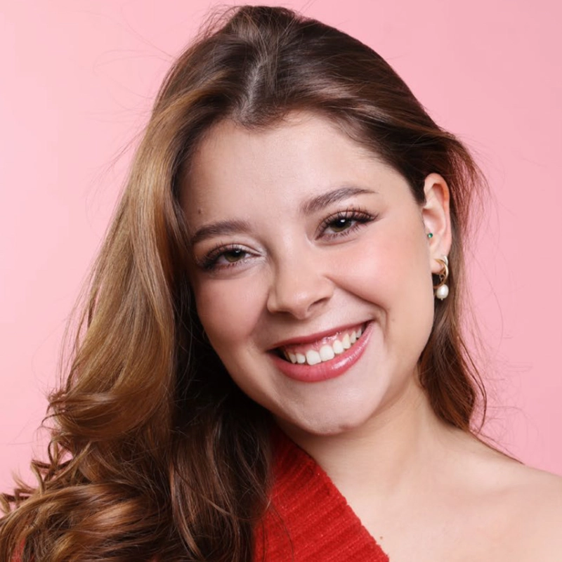 A young woman with long, wavy brown hair smiles warmly at the camera. Déa Thatcher wears subtle makeup, pearl earrings, and a red top. The solid light pink background adds a soft and cheerful atmosphere to the portrait.