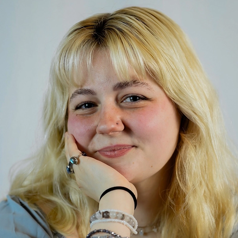 A young woman with long, wavy blonde hair and bangs smiles softly at the camera. She rests her face on her hand, wearing several rings and beaded bracelets. She has a light complexion, a nose piercing, and a black hair tie on her wrist, with a plain background.