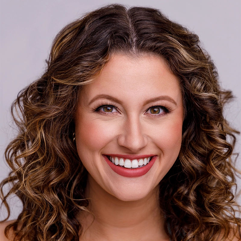 Jenna Willet, a woman with long, brown, curly hair and hazel eyes, smiles warmly at the camera. She has even makeup with pink lipstick and wears small hoop earrings. The soft, light gray background keeps the focus on her face.