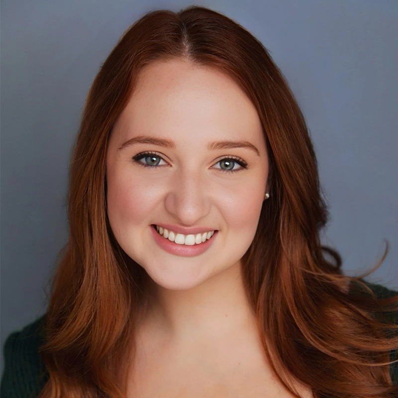 A young woman with long, wavy auburn hair smiles at the camera. She has fair skin, light-colored eyes, and is wearing natural makeup. She is dressed in a dark top and poses in front of a plain, soft blue background.