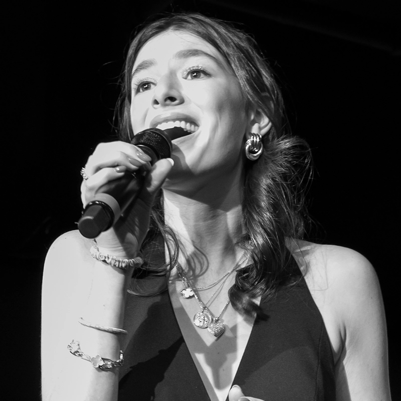 A woman with wavy hair, wearing earrings, necklaces, and a sleeveless top, sings into a microphone on stage. She smiles brightly, lit by stage lights, with one hand holding the mic and the other partly visible. The image is in black and white.