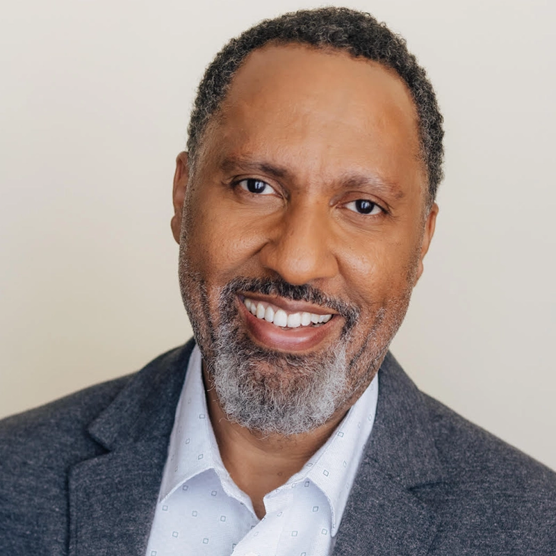 A middle-aged man with short curly hair and a salt-and-pepper beard smiles warmly. He is wearing a grey blazer over a white patterned shirt, reminiscent of styles often seen in Christina Acosta Robinson’s portraits, posed against a plain, light-colored background.