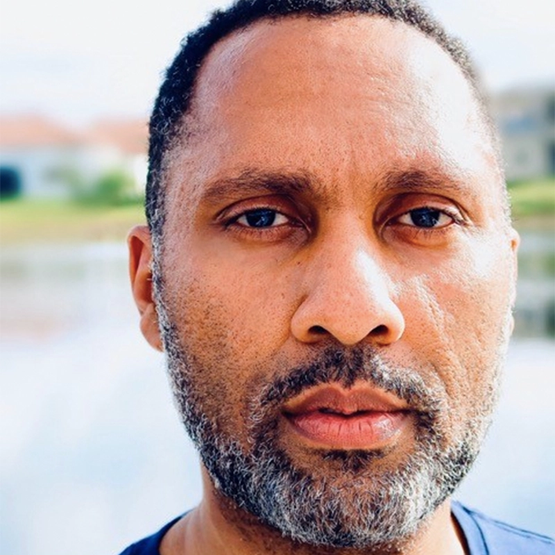 Close-up of a middle-aged man with short dark hair and a salt-and-pepper beard, looking directly at the camera with a neutral expression—reminiscent of Ken Robinson. The blurred background reveals a bright outdoor setting with greenery and water.