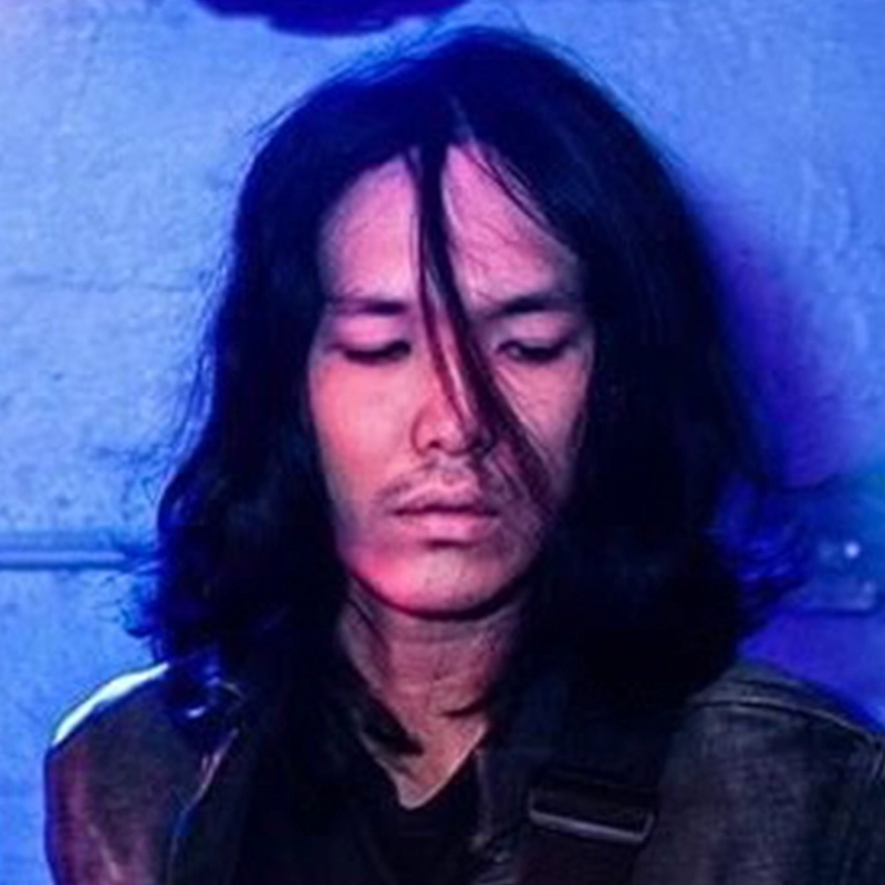 Kenji Tokunaga, with long black hair and a mustache, looks downward with a serious expression. Wearing a dark jacket and shirt, he stands against a wall lit by blue and purple lighting, his hair partially covering his face.