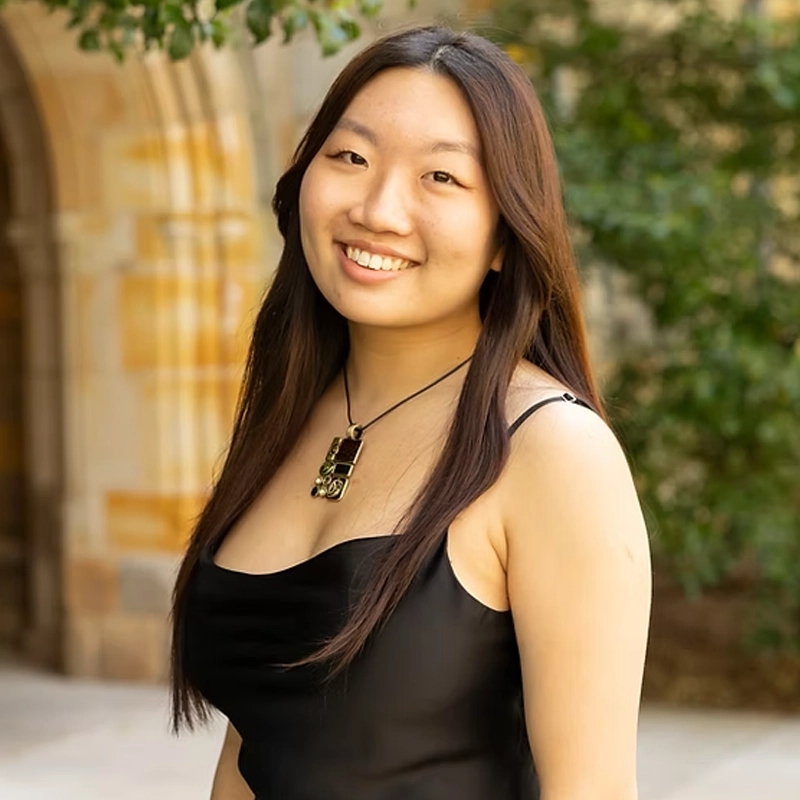 A young woman with long brown hair wears a sleeveless black dress and a black necklace. She smiles at the camera, standing outdoors near a stone building with arches and green foliage in the background.
