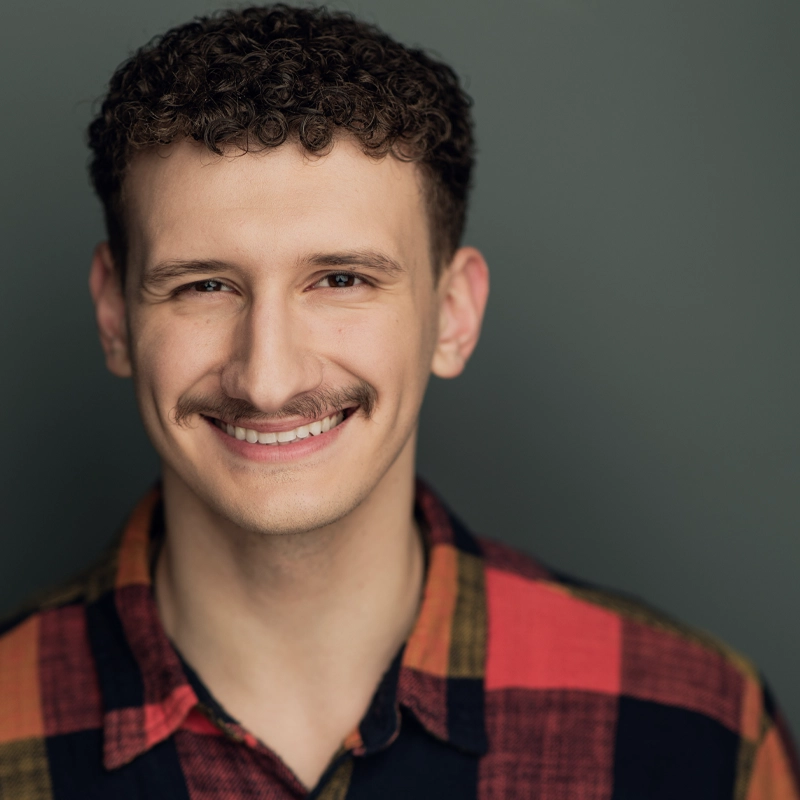 A young man with short curly brown hair and a mustache smiles warmly. He wears a plaid shirt with red, yellow, and black. The soft lighting and plain muted greenish-gray background give Konstantin Kipshidze a friendly, approachable feel.