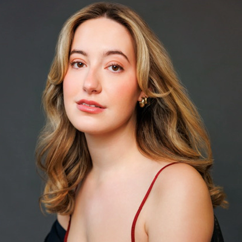 A young woman with wavy blonde hair and fair skin poses in front of a dark gray background. She wears a black top with thin red straps and gold hoop earrings. She gazes at the camera with a soft, gentle expression and natural makeup.