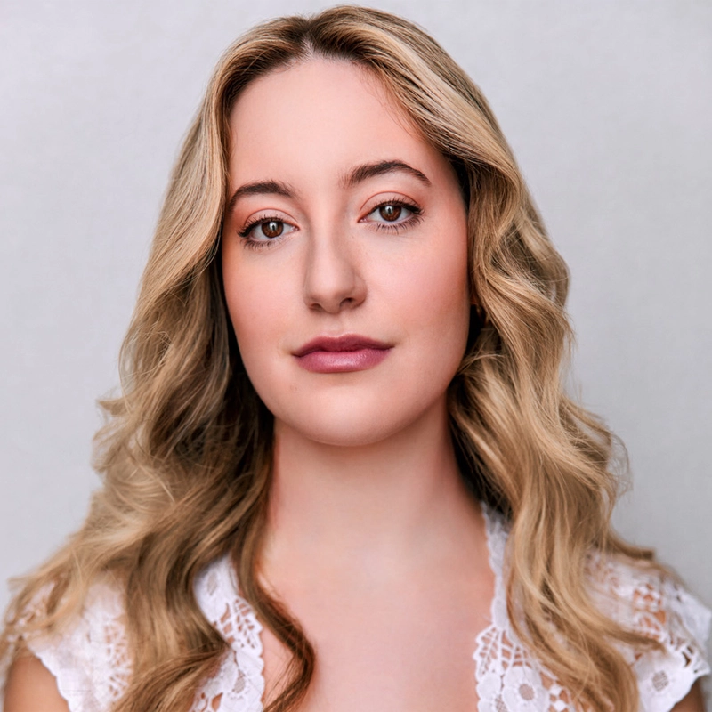 A woman with long, wavy blonde hair and fair skin looks directly at the camera. She has soft makeup with pink lips and is wearing a white lace top. The background is plain and light-colored, creating a simple, elegant portrait.