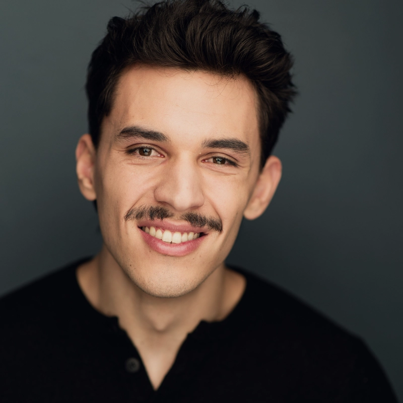 Korey Kristensen, a young man with short dark hair and a mustache, smiles at the camera. He wears a black shirt and is posed against a dark, plain background, giving the portrait a professional and friendly appearance.