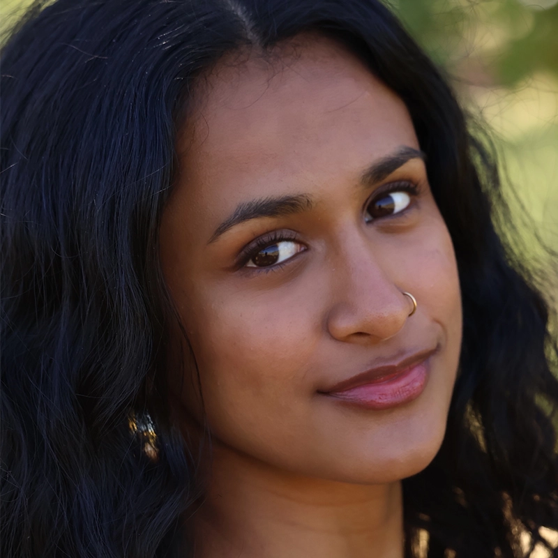 Addison Zacharias, with long, wavy black hair and medium brown skin, looks at the camera with a gentle smile. They have a gold nose ring and subtle makeup, standing outdoors with blurred greenery in the background.