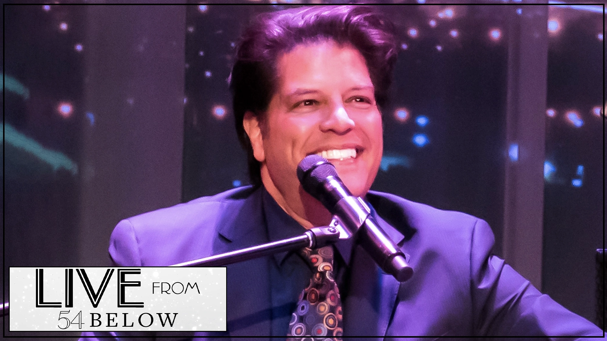 A man in a suit and patterned tie, possibly George Bugatti, smiles broadly while singing or speaking into a microphone on stage, surrounded by soft, colorful lights. The bottom left corner features a 