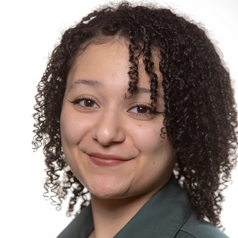 A young woman with curly brown hair smiles gently. She wears natural makeup with winged eyeliner and a dark green collared top. The white background and bright lighting create a clear, professional look. Her head is slightly tilted, and her expression is warm—capturing the approachable presence of Eden Lakey-Wright.