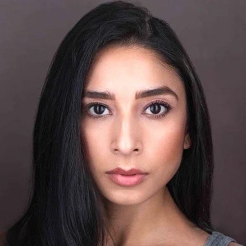 A young woman with long, straight black hair and warm-toned skin looks directly at the camera. She has dark, expressive eyes, defined eyebrows, and neutral makeup. The background is grayish-brown; her calm, serious expression suggests Violet Tinnirello.