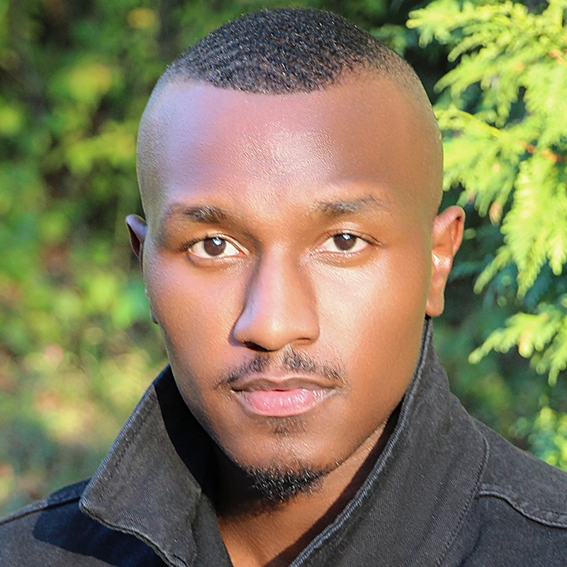 A young Black man with short hair and a trimmed beard looks directly at the camera. Wearing a dark jacket, Lazarus Rashid stands against a backdrop of green foliage and sunlight, creating a natural outdoor setting.