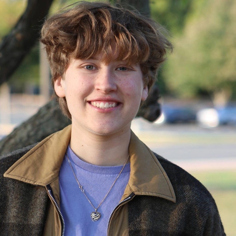 A person with short, tousled brown hair and light skin is smiling outdoors. They wear a brown plaid jacket with a tan collar over a lavender shirt and a silver necklace. The background is blurred trees and a street, giving a natural, sunny setting.