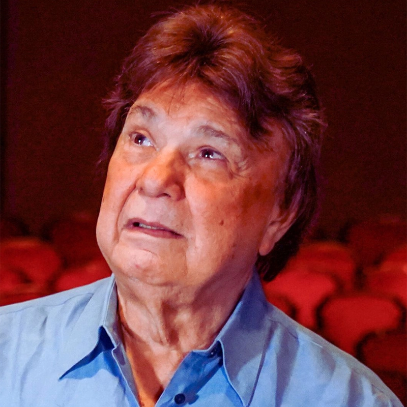 An older man with medium-length brown hair and wearing a light blue shirt looks slightly upward with a thoughtful expression. The blurred background of red theater seats suggests Rafael Lebrón is deep in contemplation.
