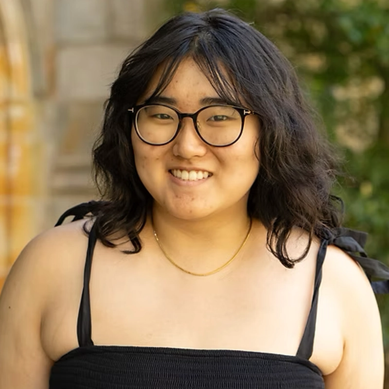 A person with wavy black hair, glasses, and a gold necklace smiles at the camera. They wear a black sleeveless top with shoulder ties. The background features blurred greenery and a stone building.