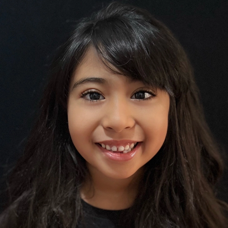 A young girl with long, dark hair and bangs smiles warmly at the camera. She has big, dark eyes, light brown skin, and is wearing a black top. The background is solid black, making her face the focus of the image.