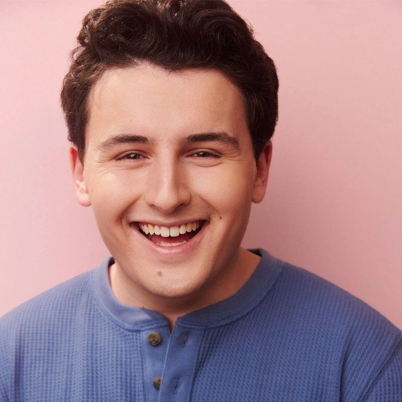 A young man, Andrew Levin, with short, dark curly hair smiles warmly. He wears a blue, buttoned Henley shirt. The soft pink background creates a bright and cheerful atmosphere, highlighting his friendly and inviting expression.