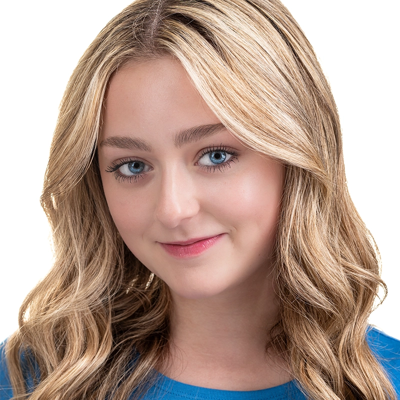 A young woman with long, wavy blonde hair and blue eyes smiles softly at the camera. She is wearing a blue top and is posed against a plain white background. Her makeup is natural, highlighting her clear skin and features.