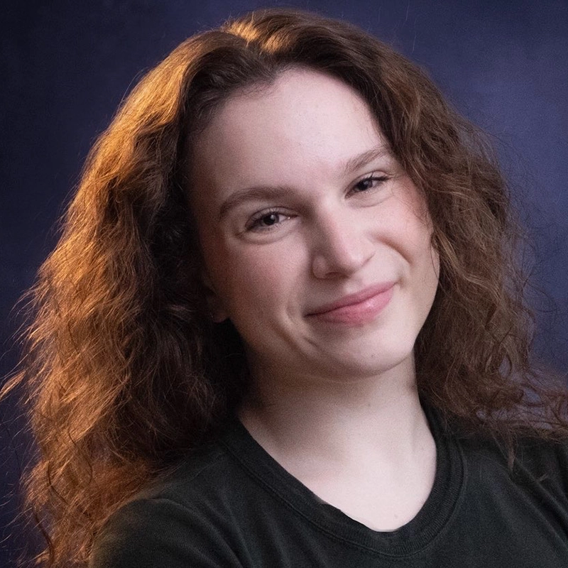 A young person with fair skin and wavy, light brown hair smiles gently. They wear a dark shirt and face the camera against a dark blue, softly lit background. The lighting highlights their hair and facial features.