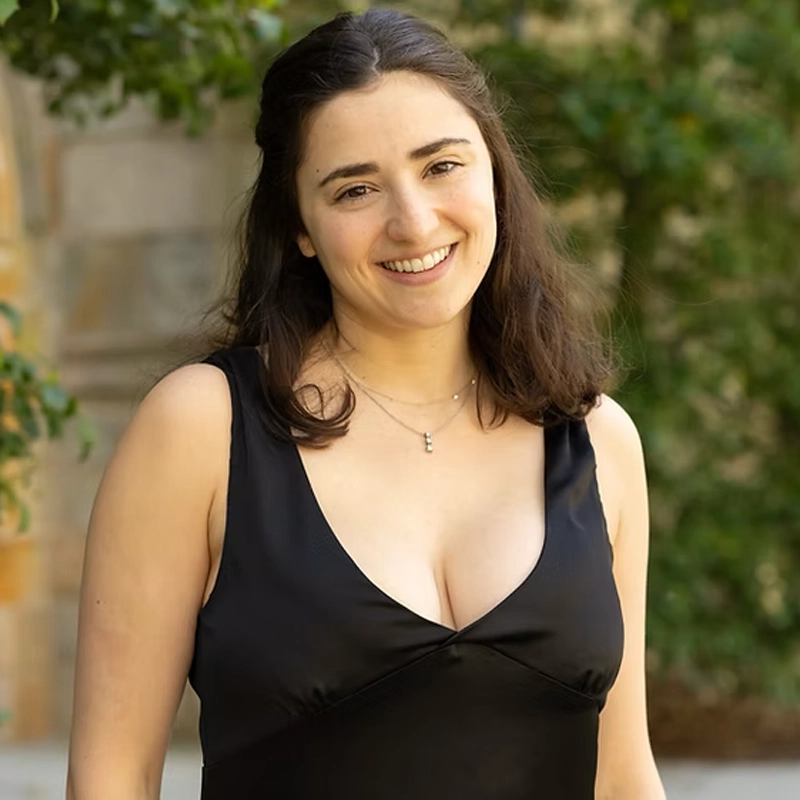 Eleanor Lockhart, a young woman with long dark hair, wears a black sleeveless dress and a small necklace as she smiles outdoors. The background is softly blurred, highlighting greenery and a stone wall.