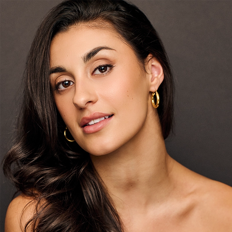 Alex Humphreys, with long, dark wavy hair, poses against a dark background. She has warm-toned skin, natural makeup, and gold hoop earrings. Smiling softly and showing her teeth, she exudes a relaxed, elegant appearance with bare shoulders.