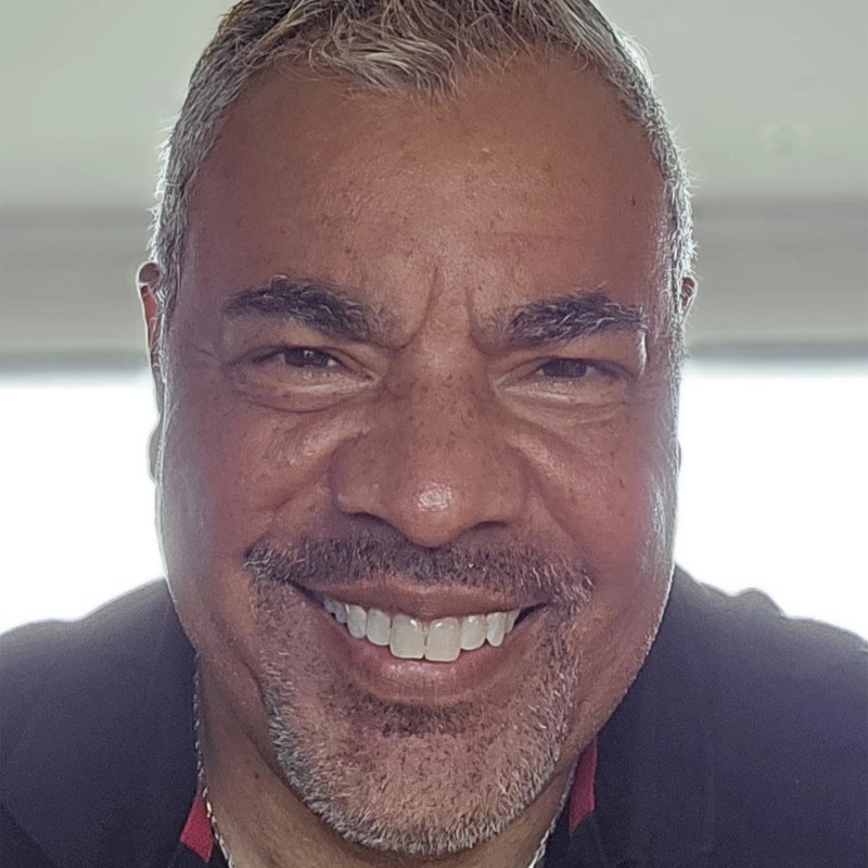 A middle-aged man with short, gray hair and a salt-and-pepper beard smiles broadly at the camera. He has warm brown skin, dark eyes, and is wearing a dark collared shirt, with natural daylight softly illuminating his face.