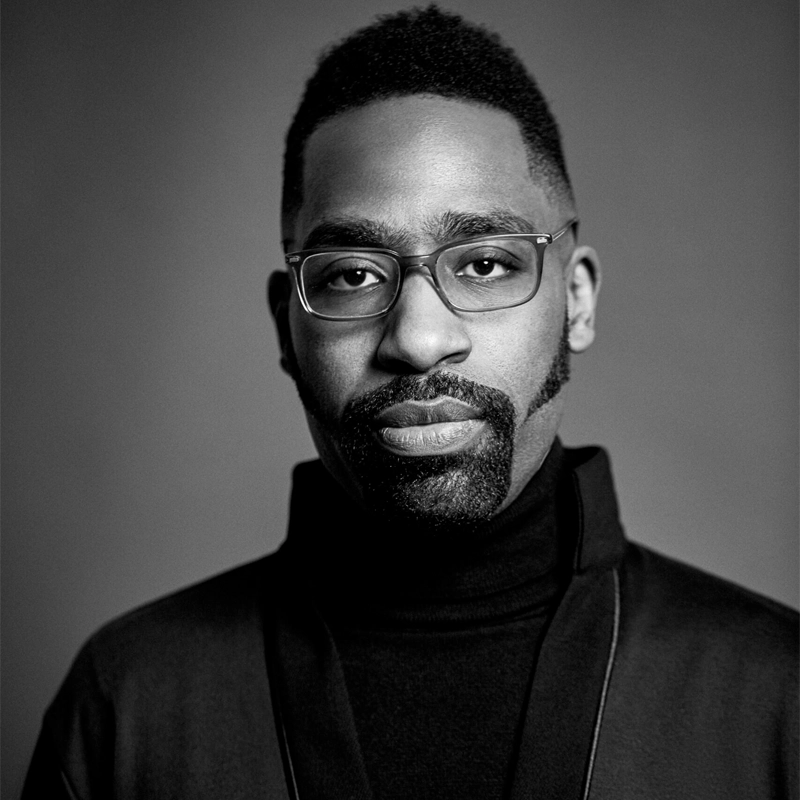 Black and white portrait of Nehemiah Luckett, a man with short hair and a neatly trimmed beard. He wears rectangular glasses, a dark turtleneck sweater, and a jacket, facing forward with a calm, neutral expression against a plain background.