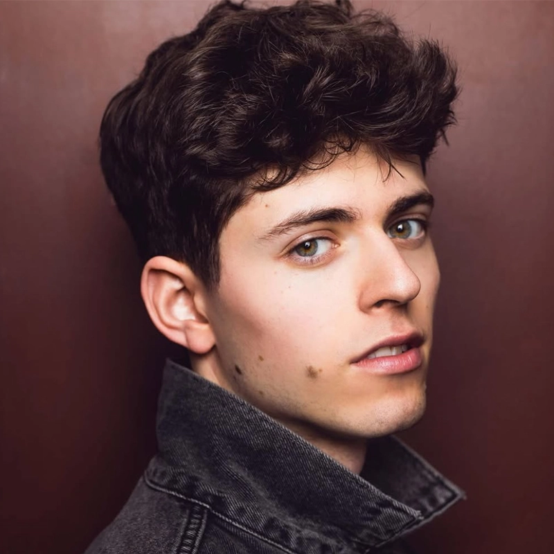 A young man with wavy brown hair and green eyes looks over his shoulder toward the camera. He has fair skin with a mole on his cheek and wears a dark denim jacket with the collar turned up, set against a plain brown background.