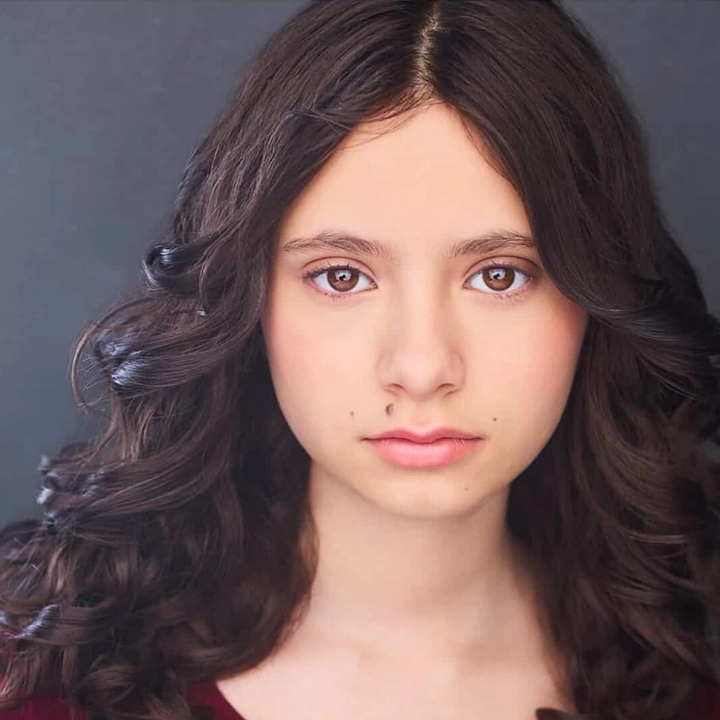 Micah Wolfensohn, a young person with long, dark, wavy hair and fair skin, stares directly at the camera with a neutral expression. They have brown eyes, two small beauty marks on their face, and stand against a plain dark gray background.