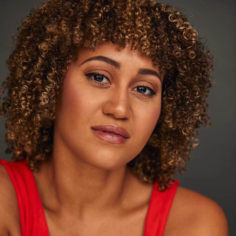 Krystal Mackie, with medium brown skin and curly light brown hair, looks confidently at the camera. She wears natural makeup with winged eyeliner and a sleeveless red top, set against a dark, neutral background.