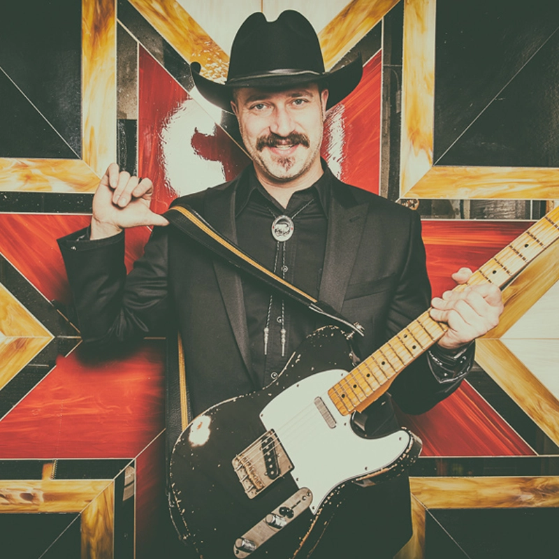 Paul Maddison, in a black cowboy hat and suit, stands smiling before a colorful geometric wall. Sporting a mustache, he holds an electric guitar with one hand while the strap rests on his shoulder.