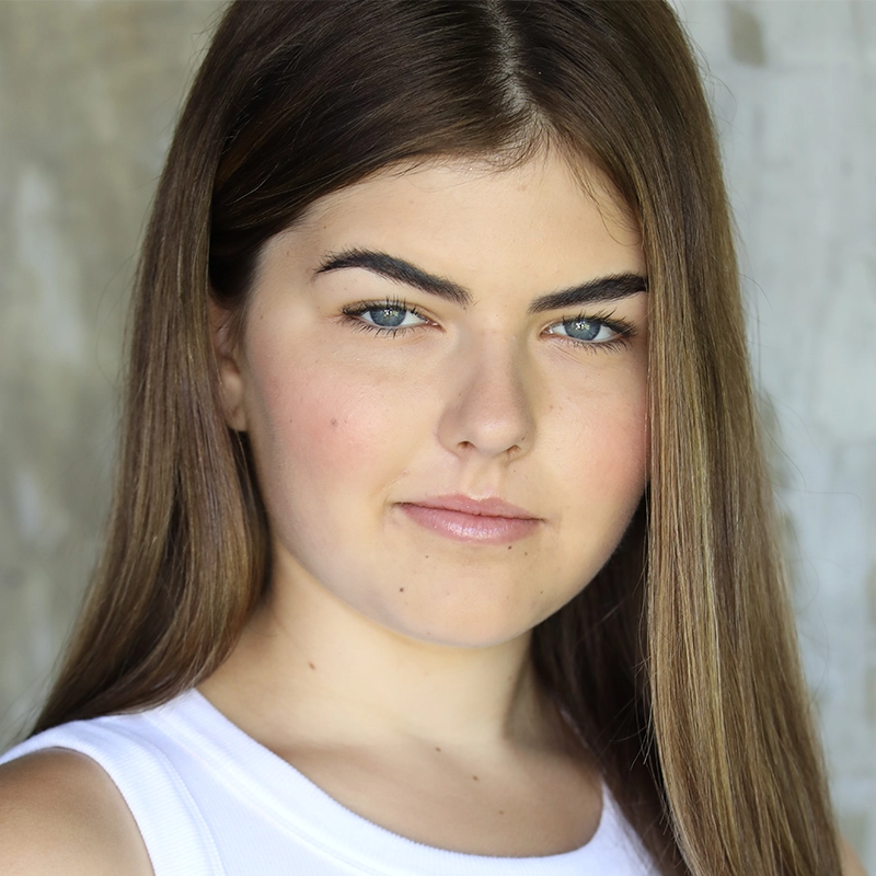 Young woman, Sara Ross Avery, with straight, long brown hair and blue eyes looks at the camera with a neutral expression. She wears a white sleeveless top. The background is blurred in soft gray and beige tones, keeping the focus on her face.