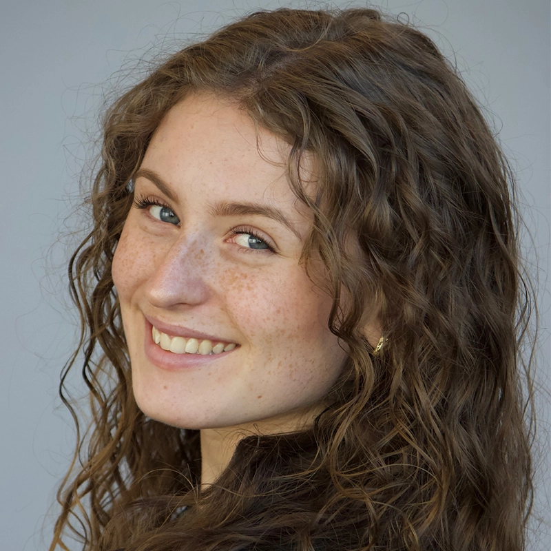 A young woman with long, wavy brown hair and freckles smiles at the camera. She is wearing a dark top and gold earrings. The background is a plain light gray, and she is turned slightly to the side, creating a friendly, cheerful expression.