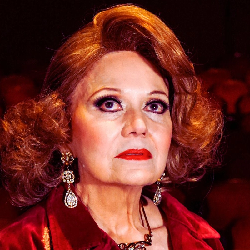 A mature woman with fair skin and voluminous auburn hair, reminiscent of Ilya Martínez, gazes upward with a serious expression. She wears dramatic makeup, red lipstick, statement earrings, and a maroon velvet outfit against a dark background.