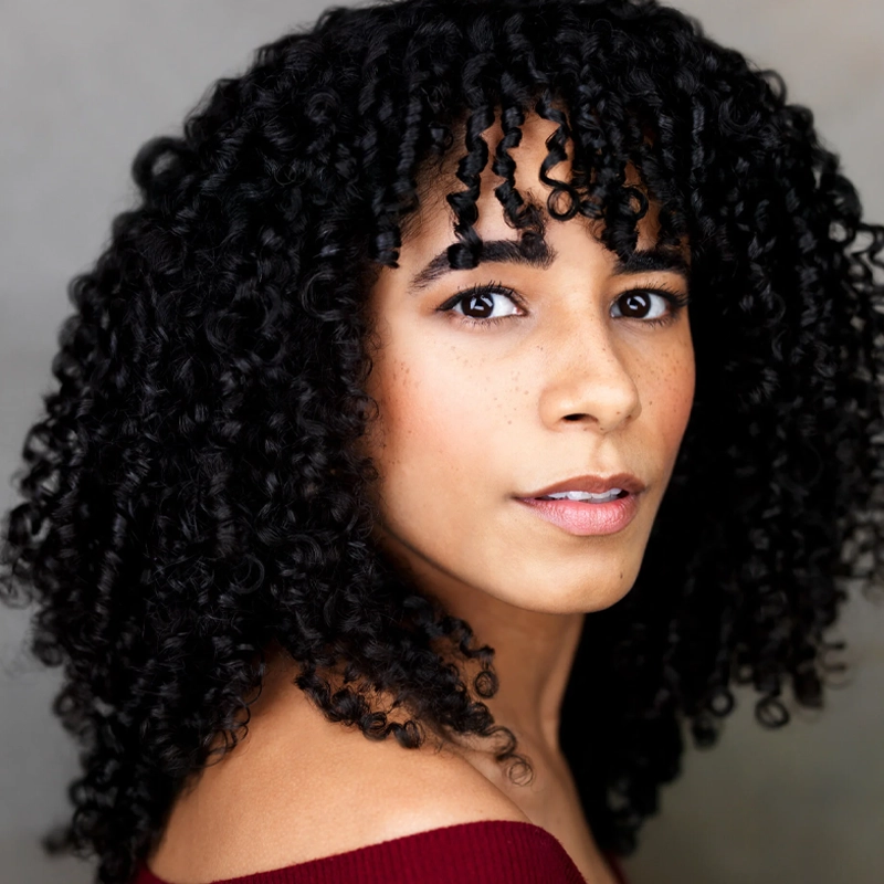 A woman with medium-dark skin and tightly curled black hair looks at the camera. She has defined eyebrows, subtle makeup, and freckles. She wears an off-the-shoulder burgundy top, and the background is softly blurred gray.