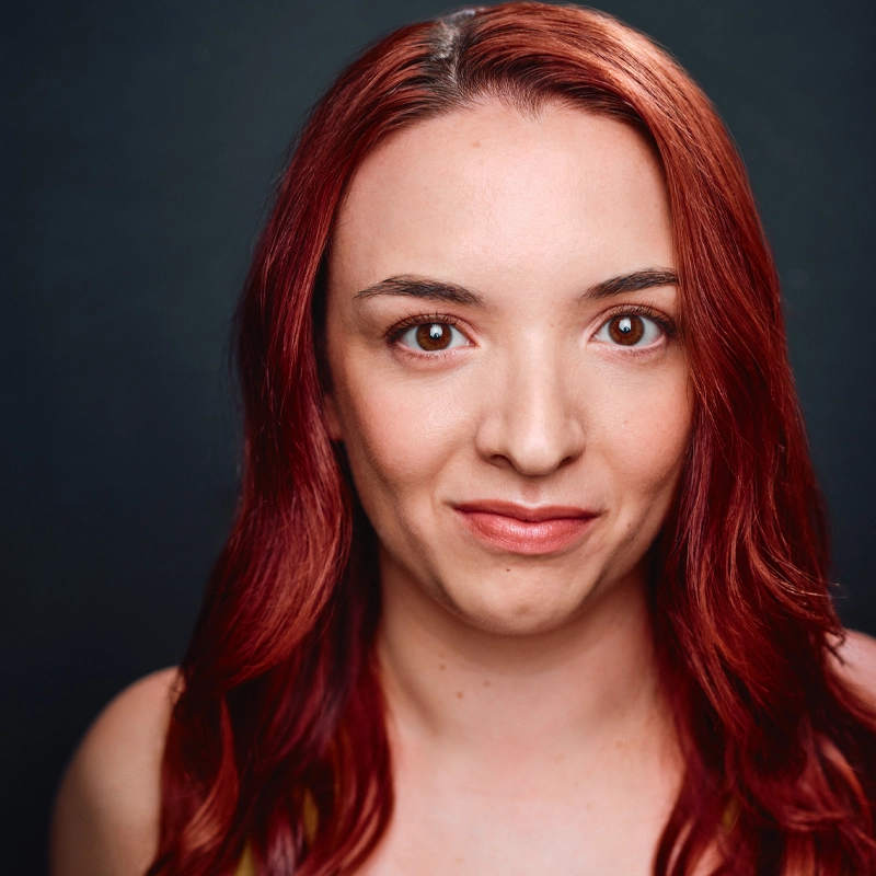 A woman with long, wavy red hair, resembling Halle Mastroberardino, smiles softly at the camera. She has fair skin, brown eyes, and wears subtle makeup. The dark, plain background draws focus to her evenly lit face.