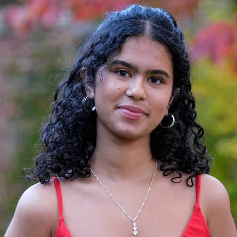 A young woman with curly dark hair, wearing hoop earrings and a delicate necklace, stands outdoors. She is dressed in a red top with thin straps. The background is blurred with autumn colors of red, yellow, and green foliage. She has a soft smile.