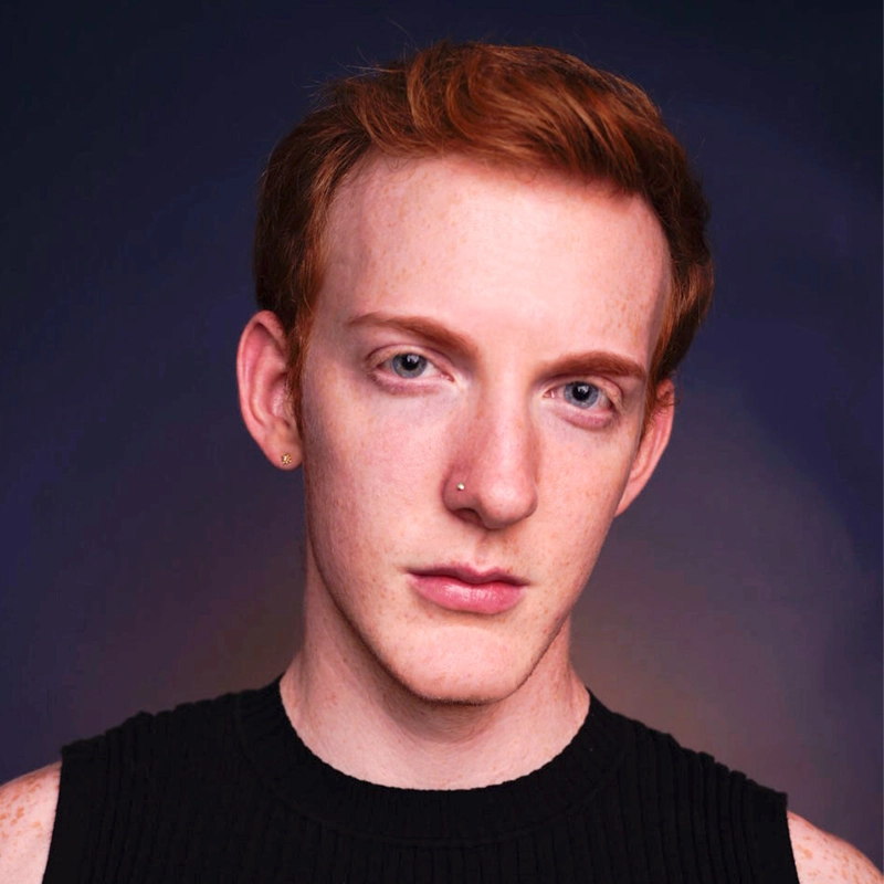 A young person with short red hair and fair skin gazes directly at the camera. They have a small nose ring and a single earring. They wear a sleeveless black top, and the background is dark with a subtle gradient.