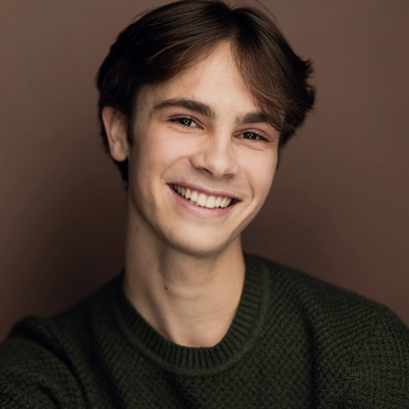 A young person with light skin, brown eyes, and brown, side-parted hair smiles warmly at the camera. They are wearing a textured, dark green sweater and are set against a plain, brown background. The lighting is soft, creating a friendly and inviting mood.
