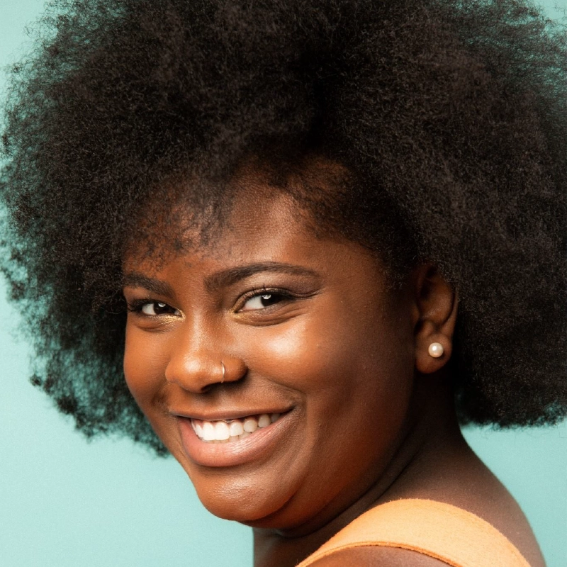 Maya J Christian, with a natural afro hairstyle, smiles warmly at the camera. She has medium-dark skin, a nose ring, and wears a single pearl earring. The background is light teal, and she is dressed in a peach-colored top.