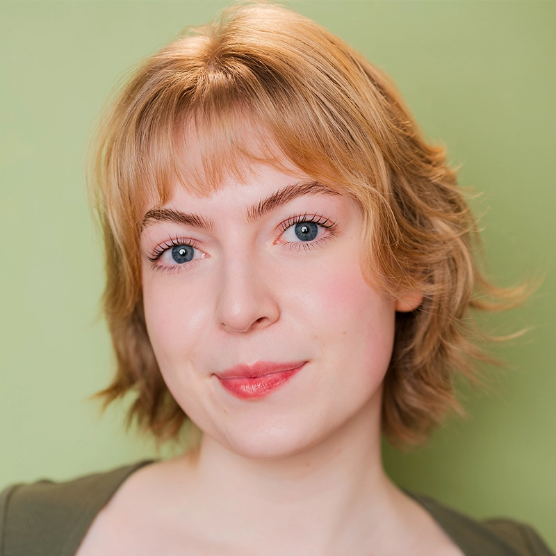 A young woman, Déa Thatcher, with short, wavy blonde hair and blue eyes smiles softly at the camera. She has fair skin, light makeup, and is wearing a green top against a plain, light green background.