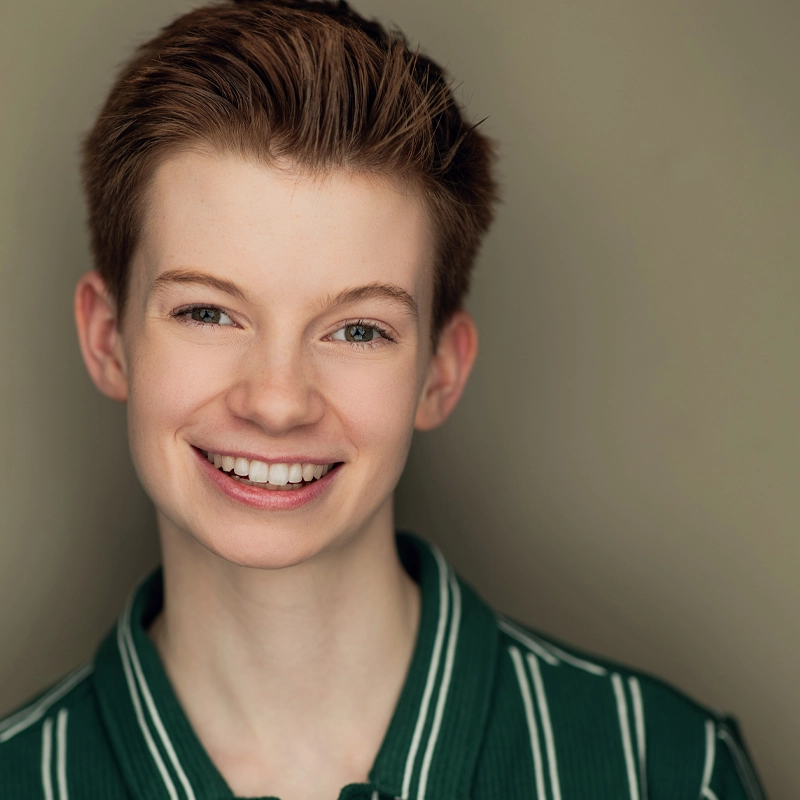 A young person with fair skin and short, light brown hair smiles warmly at the camera. Wyatt McDaniel is wearing a green collared shirt with white stripes. The background is a plain, soft beige, and the lighting is even and flattering.