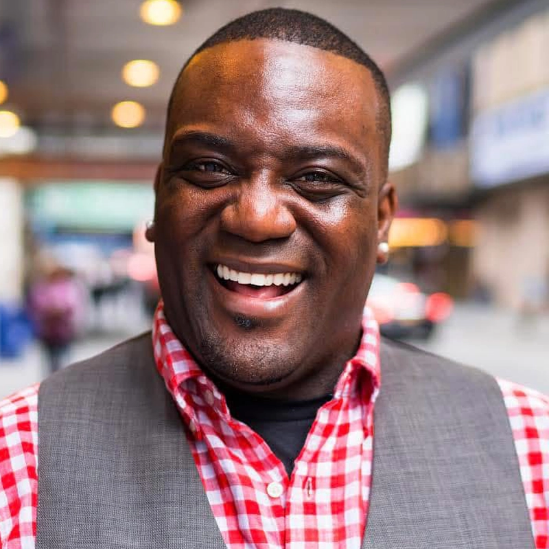 A smiling man, Peter McIntosh, wearing a red and white checkered shirt and a gray vest stands outdoors. He has short hair and earrings, with a blurred city street in the background filled with bright lights and people passing by.