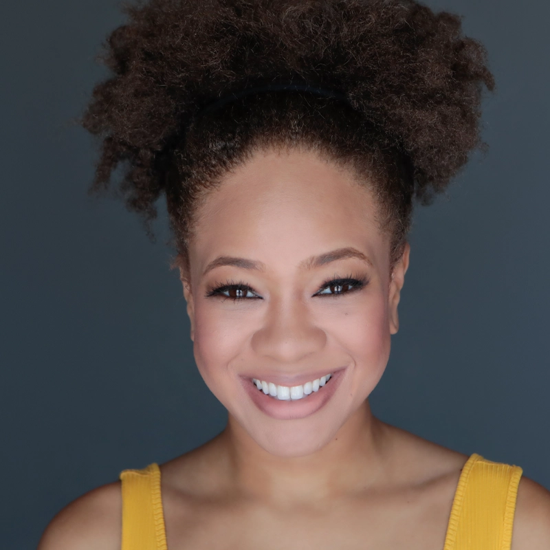 Delores McKenzie, a woman with medium brown skin and curly dark hair pulled back in a puff, smiles directly at the camera. She wears natural makeup and a sleeveless yellow top against a plain dark gray background, her expression bright and friendly.