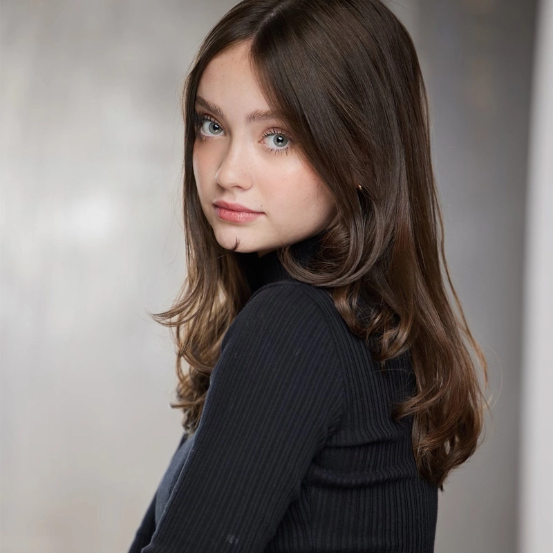 A young woman with long brown hair and blue eyes gazes over her shoulder toward the camera. She wears a black ribbed turtleneck sweater. The background is softly blurred in gray tones, drawing focus to her serene and poised expression.