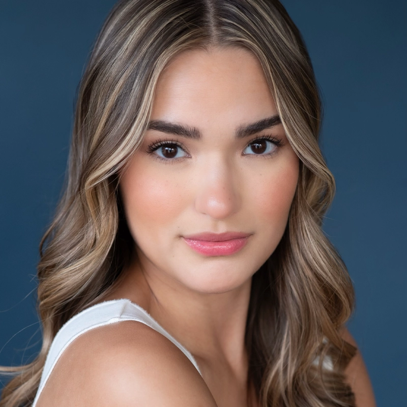 Beatriz Arevalo Medellin, a young woman with long, wavy, light brown hair and highlights, poses in front of a blue background. She has arched eyebrows, brown eyes, smooth skin, and wears natural makeup with pink lipstick and a white sleeveless top.